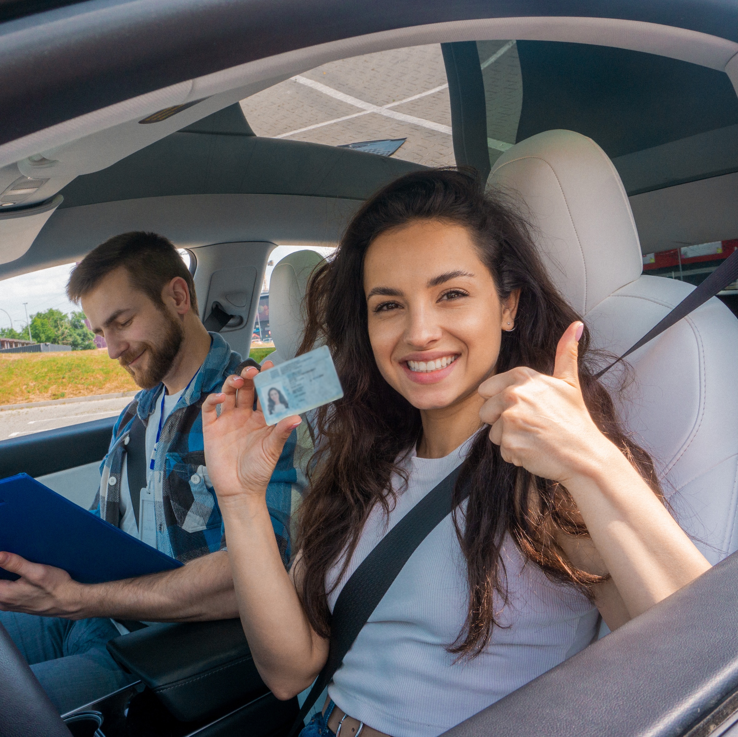 Smiling young woman with license showing thumbs up and driving school instructor with clipboard in car. Driving test, driver courses, exam concept